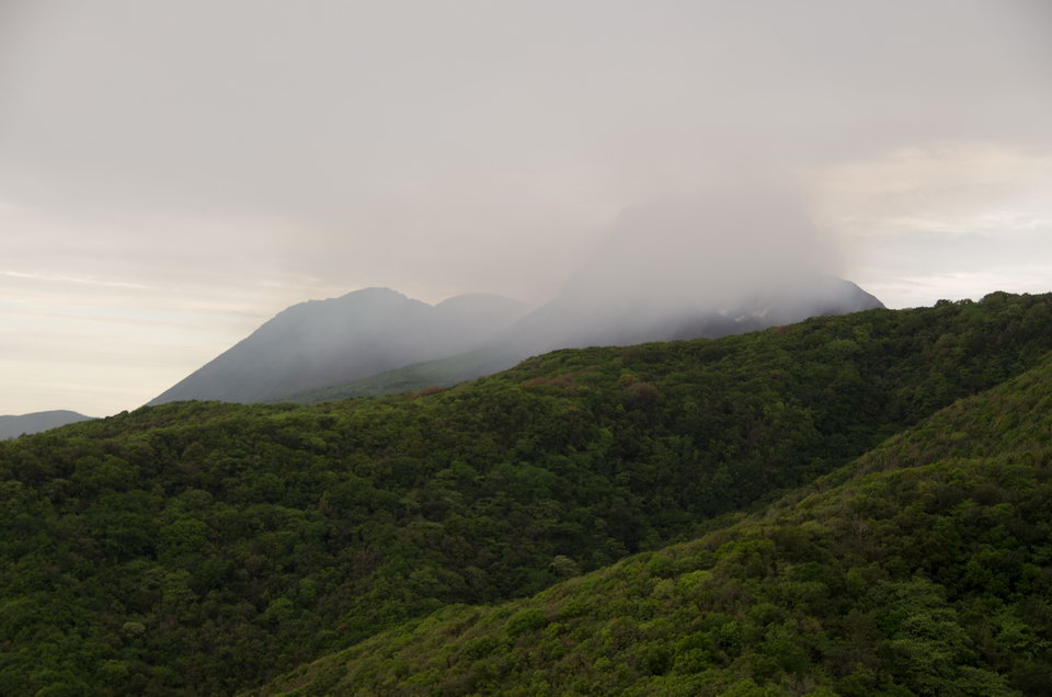 Forested hill slopes infront with the top of a volcano hidden by clouds in the background
