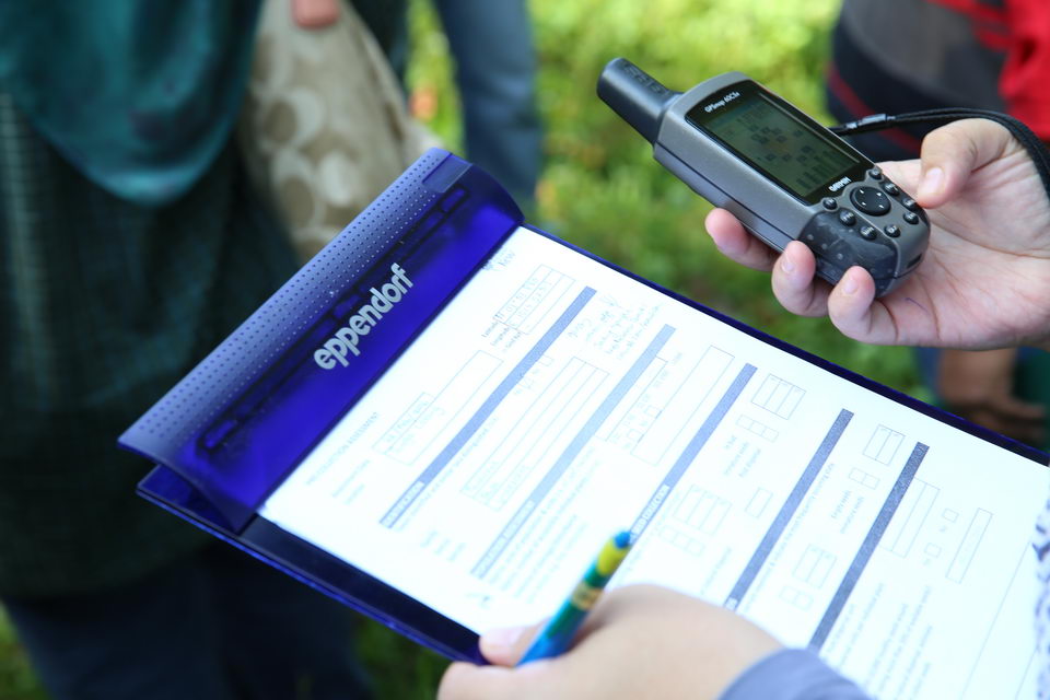 A hand holding a pen and a clipboard with a field data form on it in front of a hand holding a GPS device