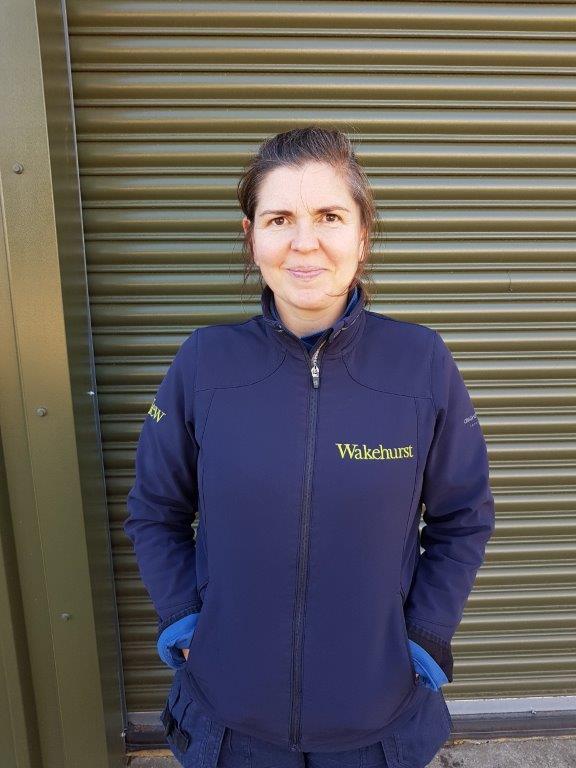 A woman stood smiling at the camera. She is wearing a navy blue jacket with Wakehurst written on it.