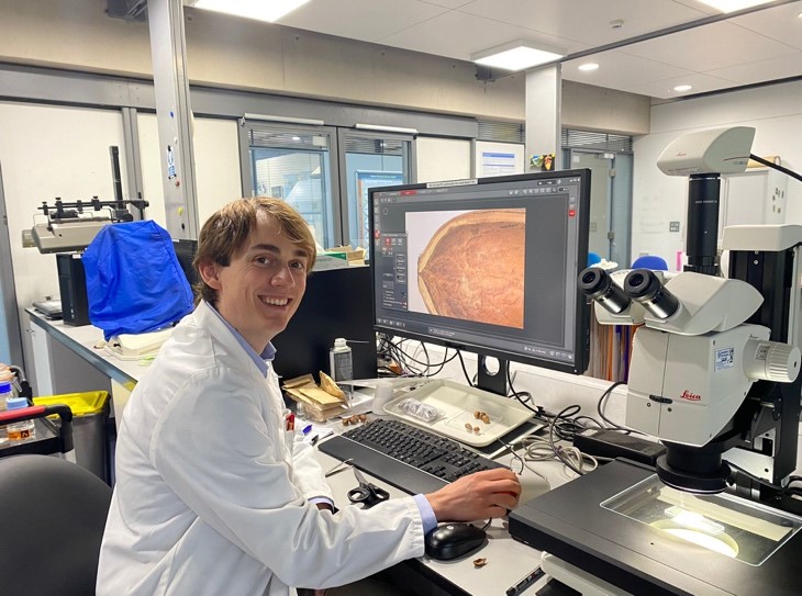 A seed scientist, sat at a laboratory bench smiling at the camera. On the bench is a computer next to a microscope, on the computer screen is an image of a seed.