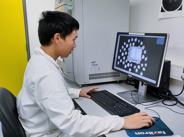 A scientist wearing a lab coat sits at a computer, with the mouse in his hand and he is focusing on the screen. On the monitor is an x-ray image showing many full tear drop shaped seeds