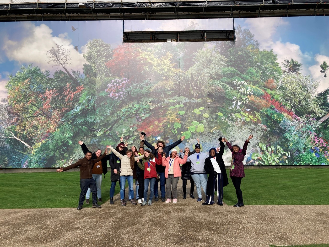 A group of people stand in front of a massive piece of outside botanical art. Some people have their hands in the air, others are showing peace signs. Everyone is smiling. 
