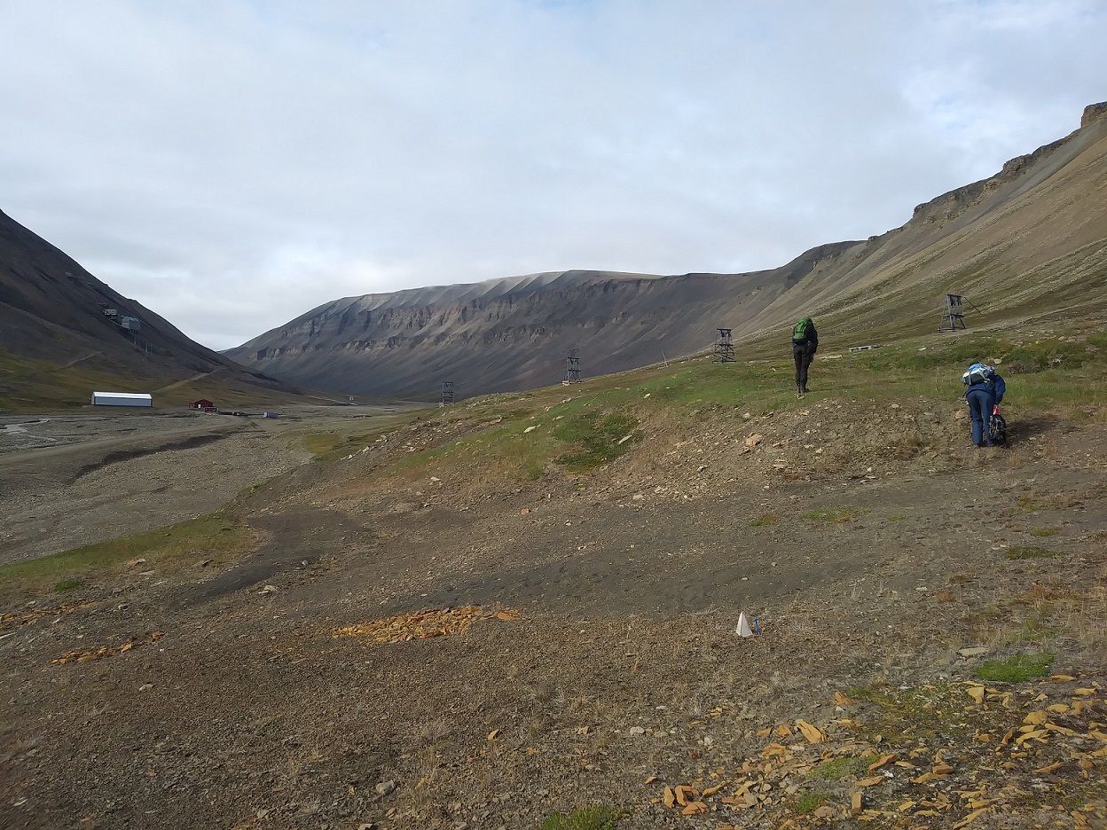 A deep glacial valley with sparse low level vegetation and a rocky substrate. Two people are visible bending down looking at vegetation