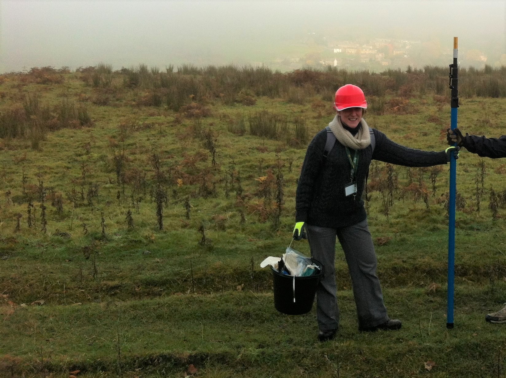 Naomi stood smiling holding a pole pruner and a bucket, wearing a hard hat on a misty hill top.