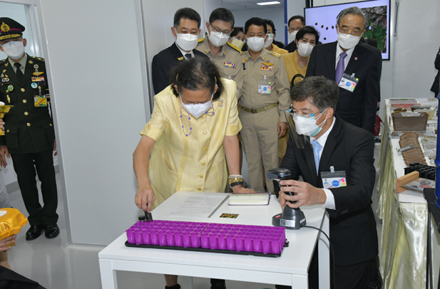 HRH Princess Maha Chakri stood behind a desk placing a seed vial into a purple container. A group of people are stood behind watching on.