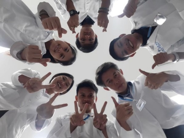 Looking up towards 6 people standing in a circle with their heads together. They are all smiling and have their thumbs up, fingers cross or in the peace sign