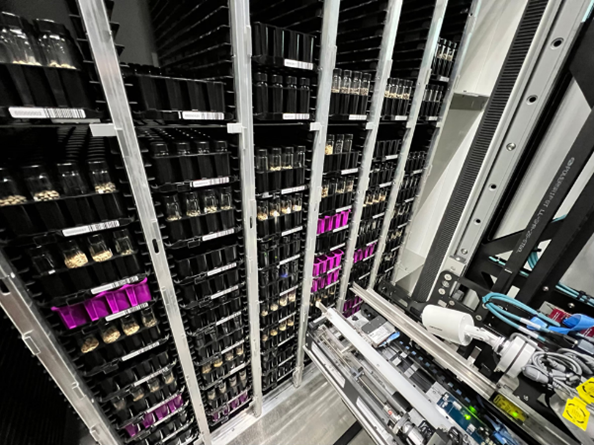 Metal racking with 6 columns of plastic trays. Some of the trays are black with glass vials sticking out from the top, whilst other trays are deeper and purple. A robotic machine is visible in front of the racking