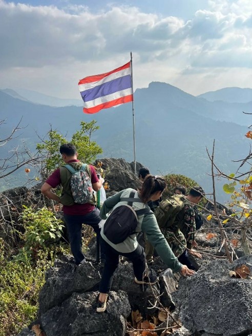 A group of three people scrambling over rocks. There is a flag pole on the edge with the Thai flag flying. There are mountains in the background.
