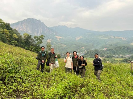 A row of six people stood smiling at the camera. They are stood amongst shrubs on a valley side with mountains in the background.