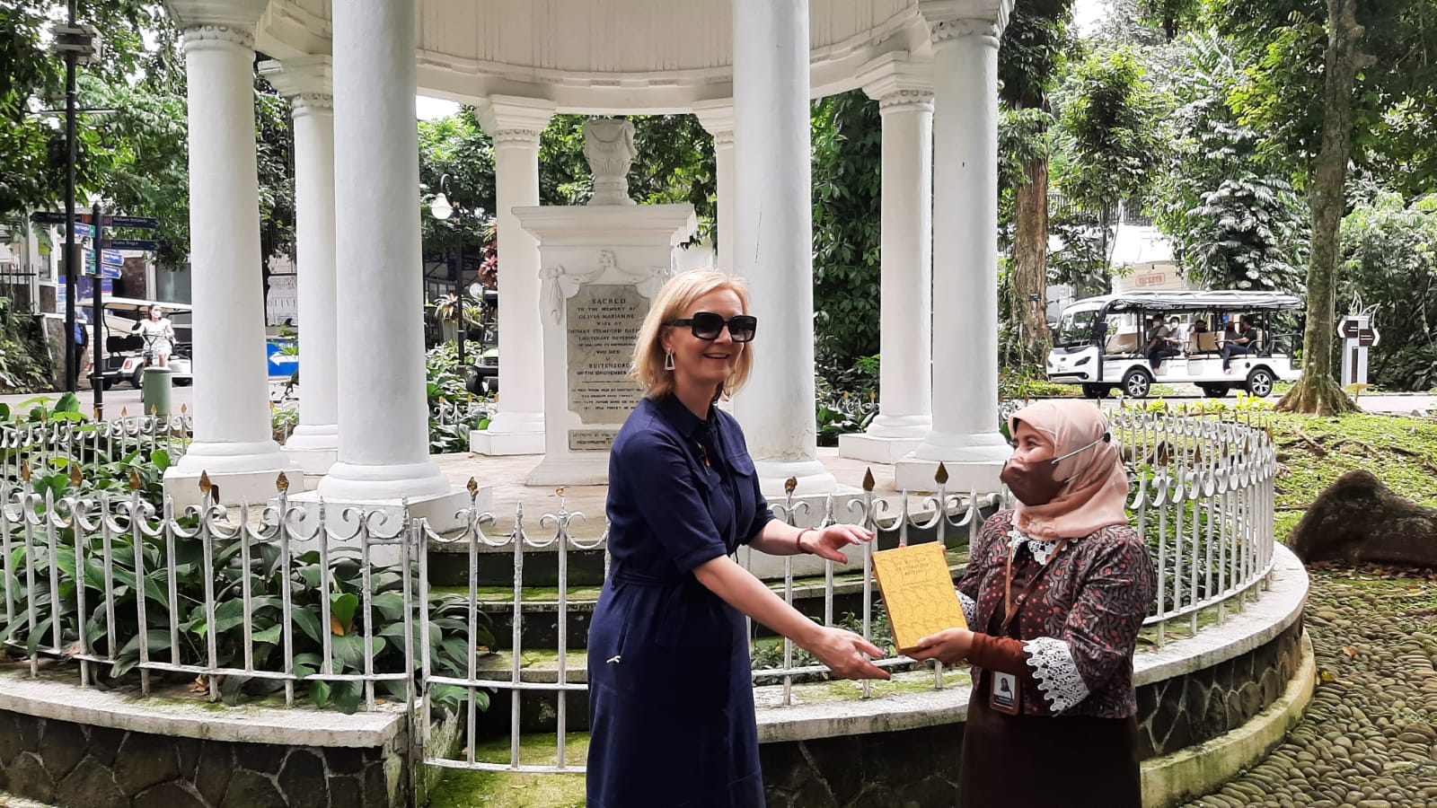 Dr Dian Latifah stood next to Liz Truss presenting her with a book. They are stood in front of a round memorial building of 8 large white pillars with a central plinth with a plaque on.